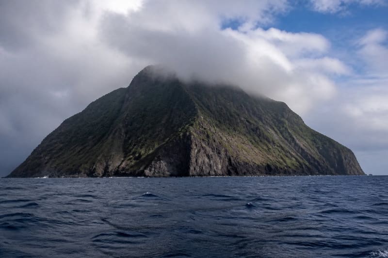 Saba from the sea: one steep volcanic island rising out of deep water, cliffs straight down, no beach, cloud on the summit.