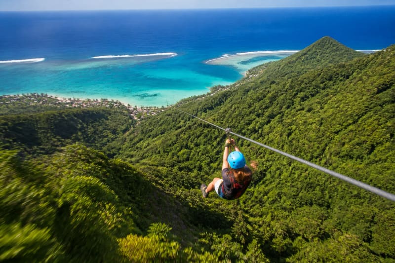 A rider on a long zipline cable running down a steep green mountainside above a wide lagoon.