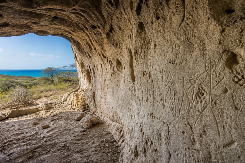 Shallow Amerindian rock carvings on the pale wall of a limestone cave, daylight coming in from the entrance.