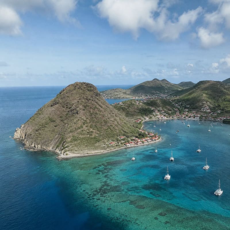 The Pain de Sucre anchorage at Les Saintes from above, yachts in clear water beside a rounded headland.