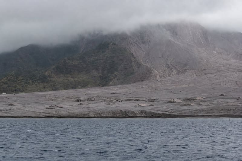 Plymouth, Montserrat, half buried in grey ash, seen from the water with the volcano behind it.