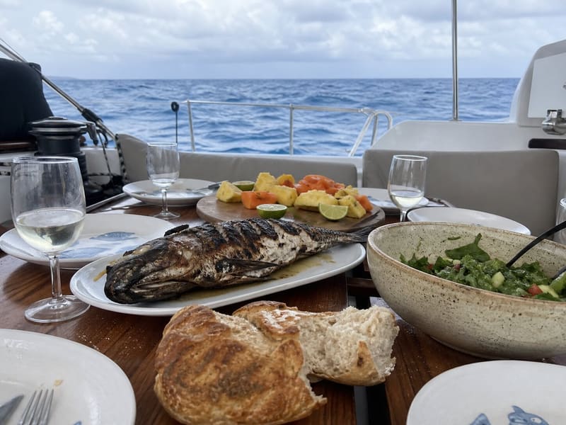 A meal on the cockpit table: whole grilled fish, bread, cut fruit and glasses of wine, the horizon just behind.