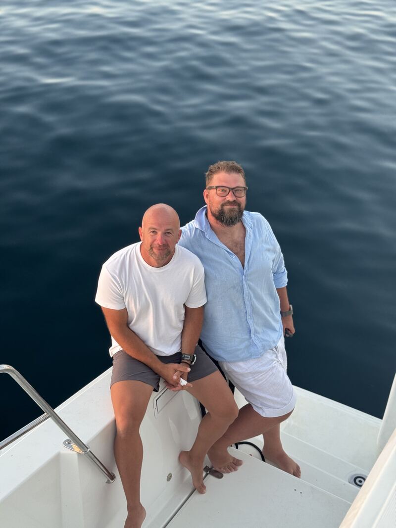 The host and the skipper sitting together on the stern step of a boat, bare feet on the deck, deep blue water behind them.
