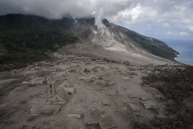 Looking down from a helicopter on the buried town of Plymouth, rooftops half swallowed by grey volcanic ash, the steaming volcano dome behind.