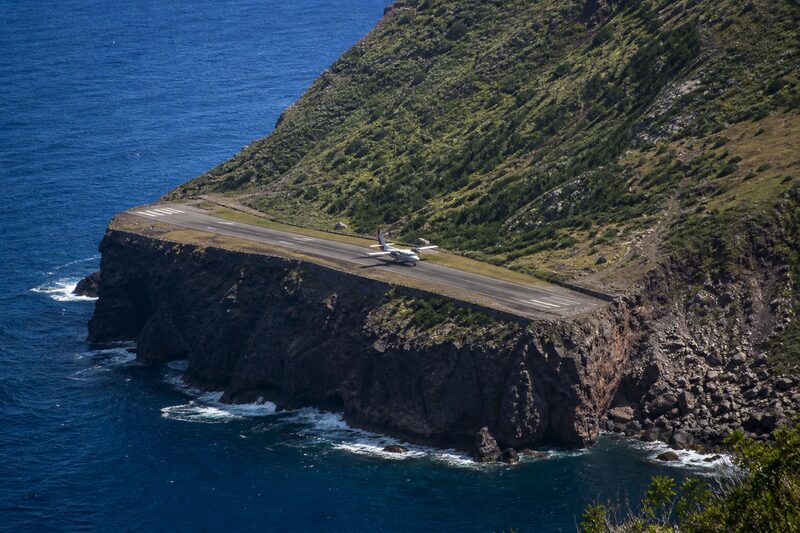 A small twin-propeller aircraft on the very short clifftop runway at Saba, black volcanic cliffs dropping into deep blue water at both ends.