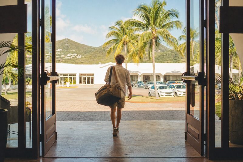 A traveller with a duffel bag walking out of a small tropical airport terminal into palms and warm afternoon light, seen from behind.