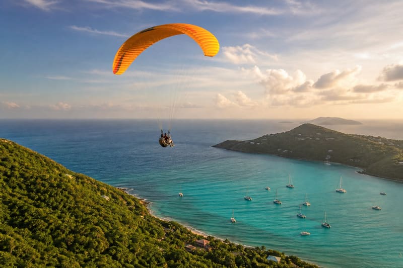 A tandem paraglider over a green Caribbean hillside, a turquoise bay and anchored boats far below.