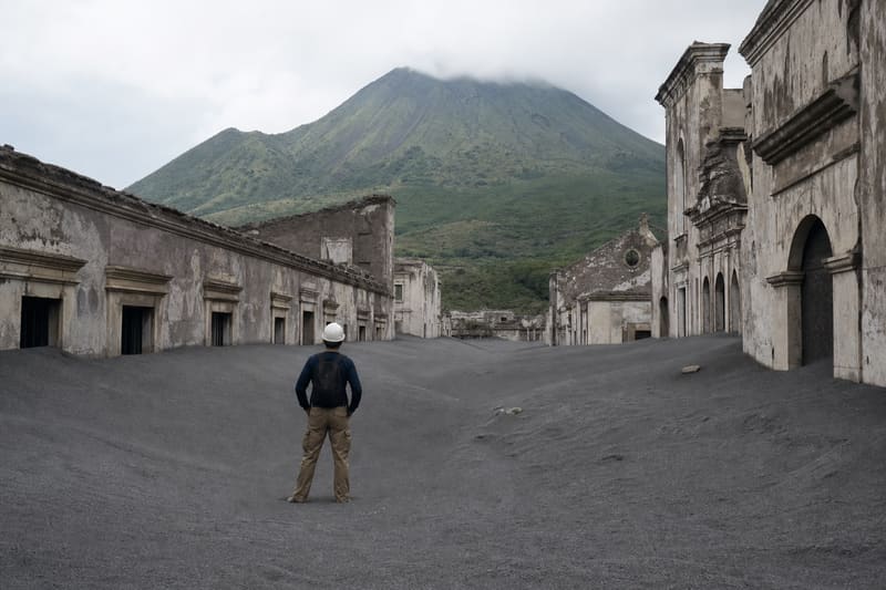 A street in an abandoned town buried to the first-floor windows in grey volcanic ash, a green volcano behind.