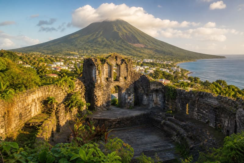 The ruined stone walls of a theatre in a Caribbean town with a large green volcano rising directly behind it.