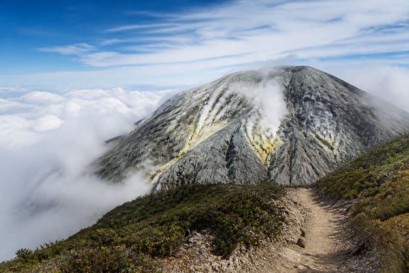 A steaming volcanic summit dome, bare grey rock and yellow sulphur, cloud below the trail.