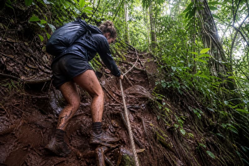 A hiker pulling up a steep muddy rainforest slope on a fixed rope.