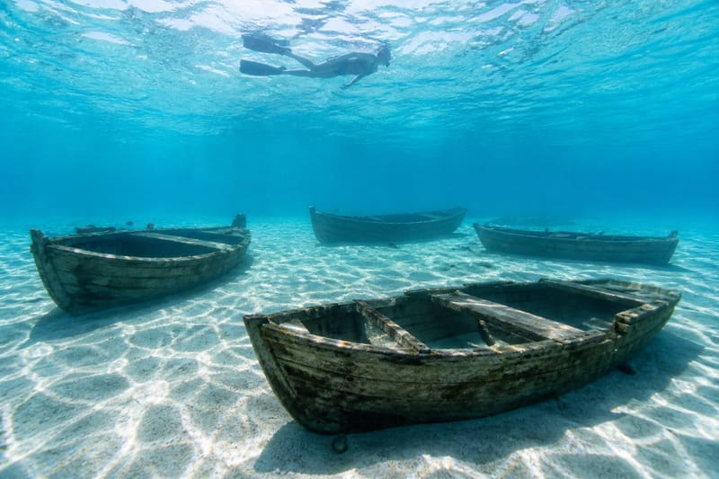 Sunken boat sculptures on pale sand in shallow turquoise water, a snorkeler at the surface above.