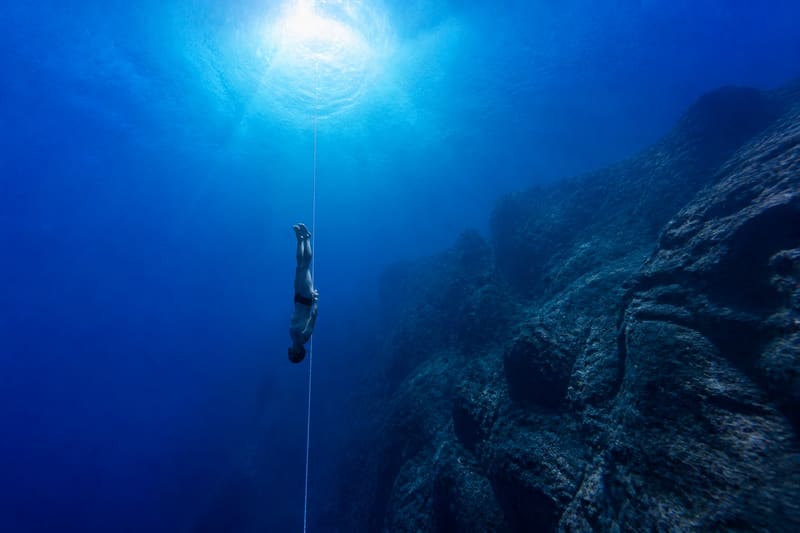 A freediver descending head-down along a weighted line, the surface a bright disc far above.