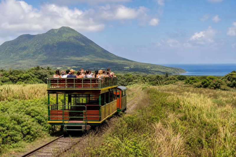 A narrow-gauge double-deck railcar running through overgrown former cane fields towards a green volcano.