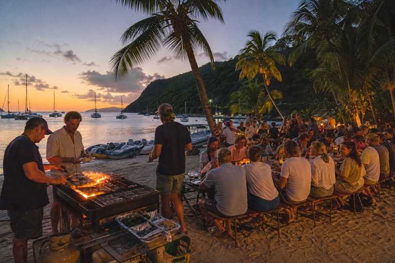A beach barbecue at dusk: grills and trestle tables under palms, dinghies pulled up on the sand.